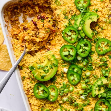 A baked casserole topped with crushed chips, sliced jalapeños, avocado, and green onions in a white dish. A serving has been scooped out with a spoon.