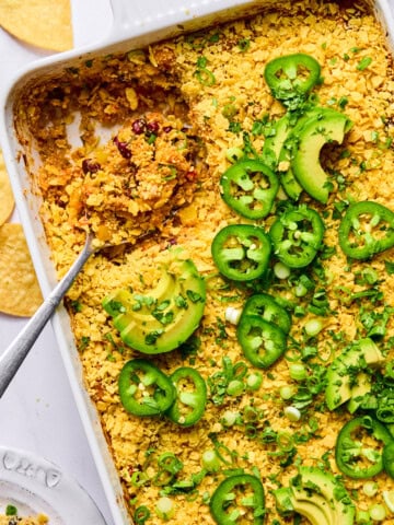 A baked casserole topped with crushed chips, sliced jalapeños, avocado, and green onions in a white dish. A serving has been scooped out with a spoon.