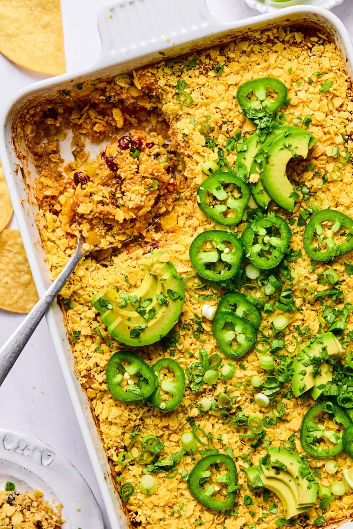 A baked casserole topped with crushed chips, sliced jalapeños, avocado, and green onions in a white dish. A serving has been scooped out with a spoon.