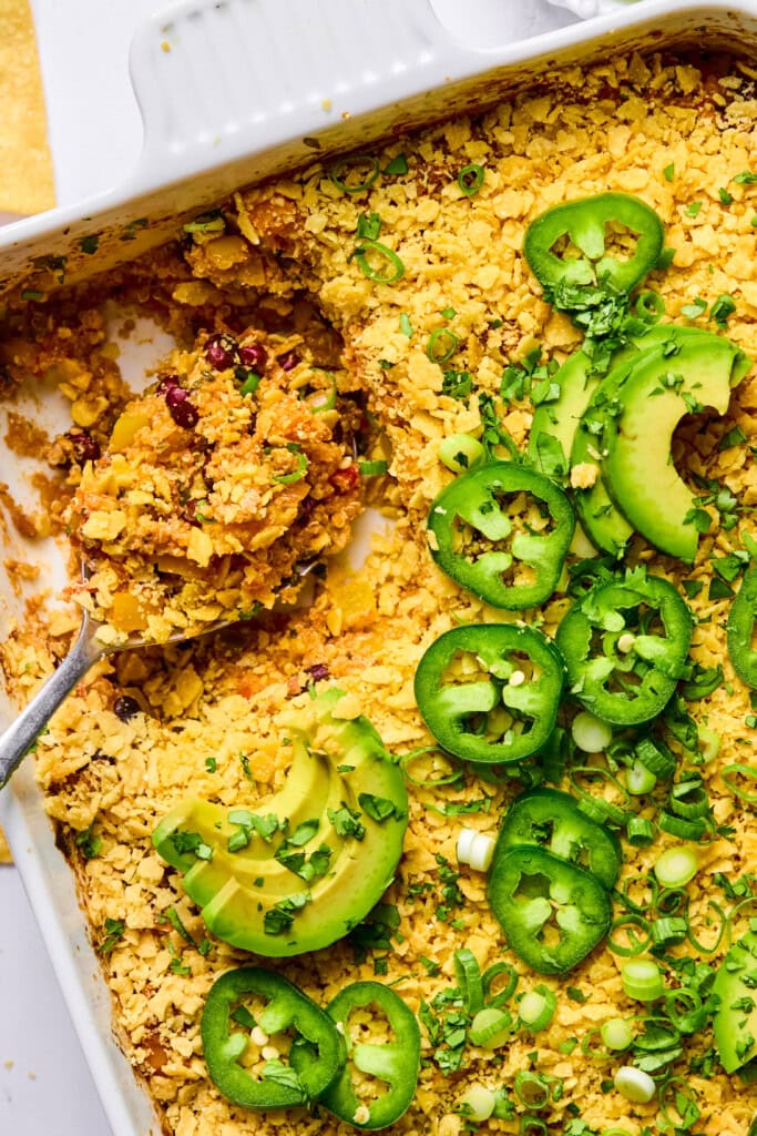 A casserole topped with sliced avocado, jalapeños, green onions, and chopped herbs. A serving has been scooped out, revealing a crumbly interior with beans and grains.