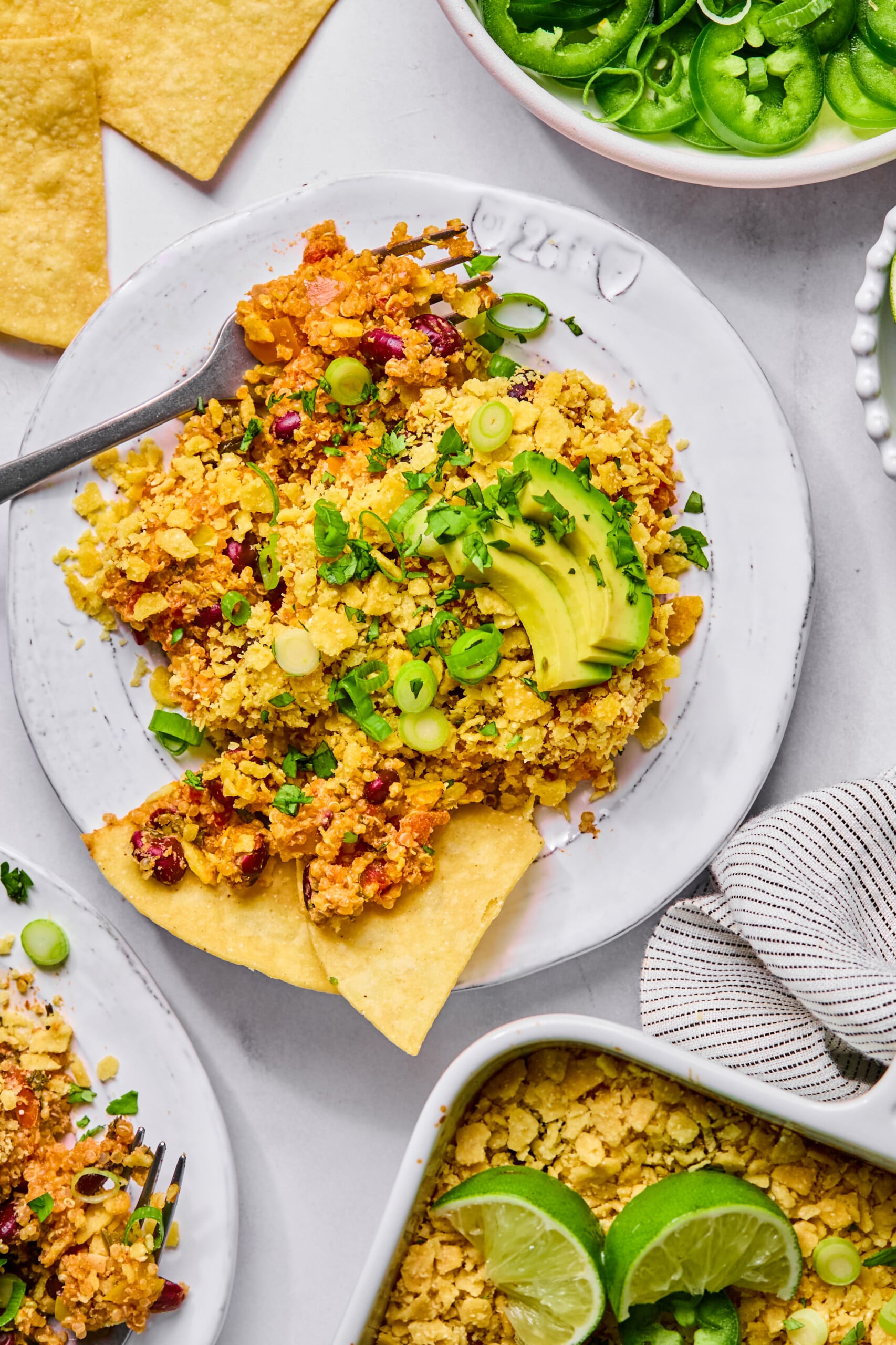 A plate of nachos topped with avocado slices, chopped scallions, herbs, and a crumbled topping, with lime wedges and sliced jalapeños on the side.