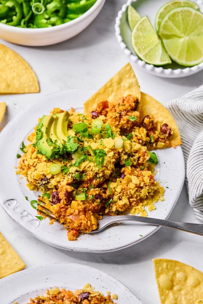 A plate of Mexican casserole topped with avocado slices, green onions, and cilantro, served with tortilla chips. Lime wedges and a bowl of green onions are in the background.