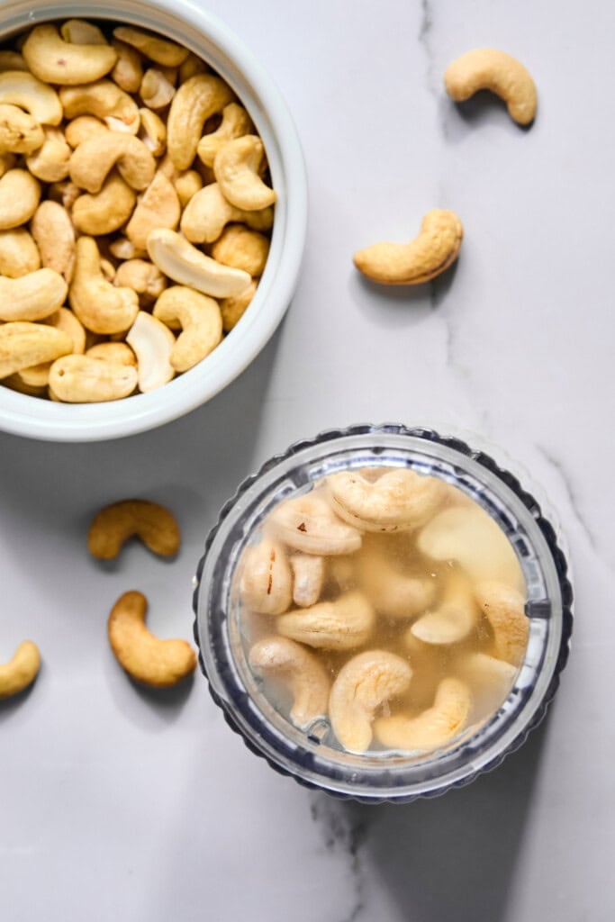 A bowl of dry cashews sits beside a blender cup filled with cashews soaking in water, on a light marble surface-just the setup you need to start making the viral tiktok watermelon ice cream recipe. Loose cashews are scattered nearby, ready for blending into a creamy base.