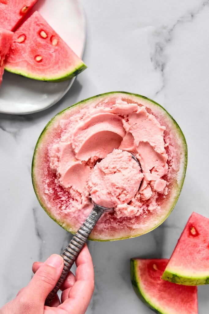 A hand scoops pink watermelon ice cream from a hollowed watermelon half, inspired by a trending tiktok watermelon ice cream recipe, with watermelon slices on a plate and the table nearby.