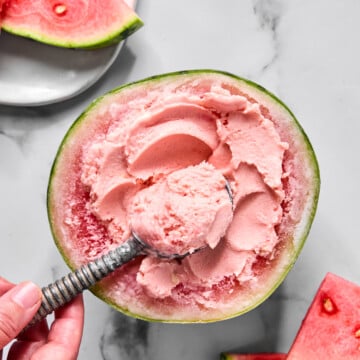 A hand scoops pink watermelon ice cream from a hollowed watermelon half, inspired by the viral TikTok watermelon ice cream recipe, with watermelon slices on a white plate and a marble surface in the background.