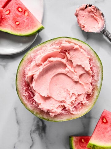 A watermelon half filled with pink ice cream, inspired by a trending TikTok watermelon ice cream recipe, is surrounded by fresh watermelon slices on a white marble surface. A metal ice cream scoop with some creamy ice cream is placed nearby.