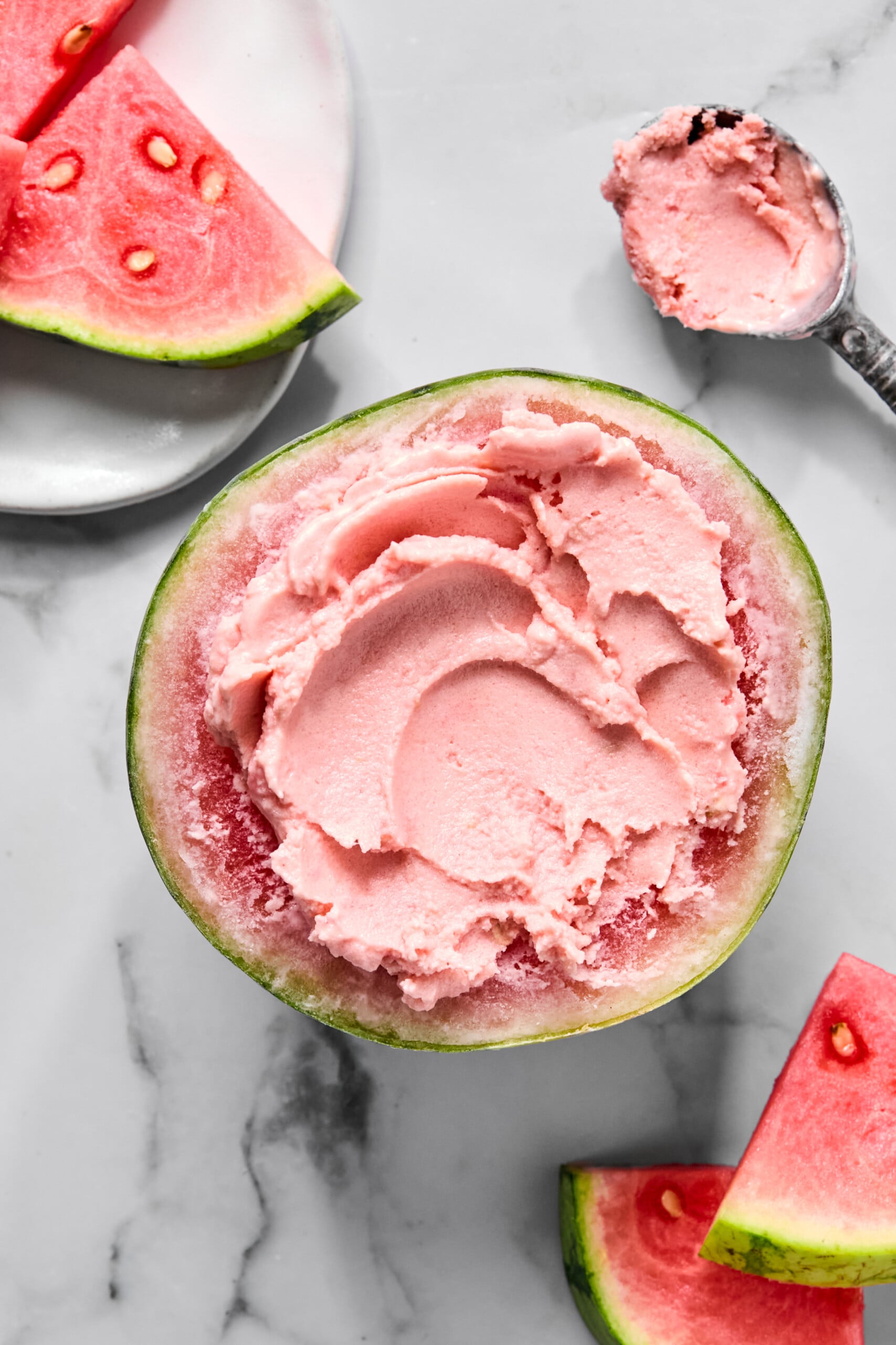 A watermelon half filled with pink ice cream, inspired by a trending TikTok watermelon ice cream recipe, is surrounded by fresh watermelon slices on a white marble surface. A metal ice cream scoop with some creamy ice cream is placed nearby.