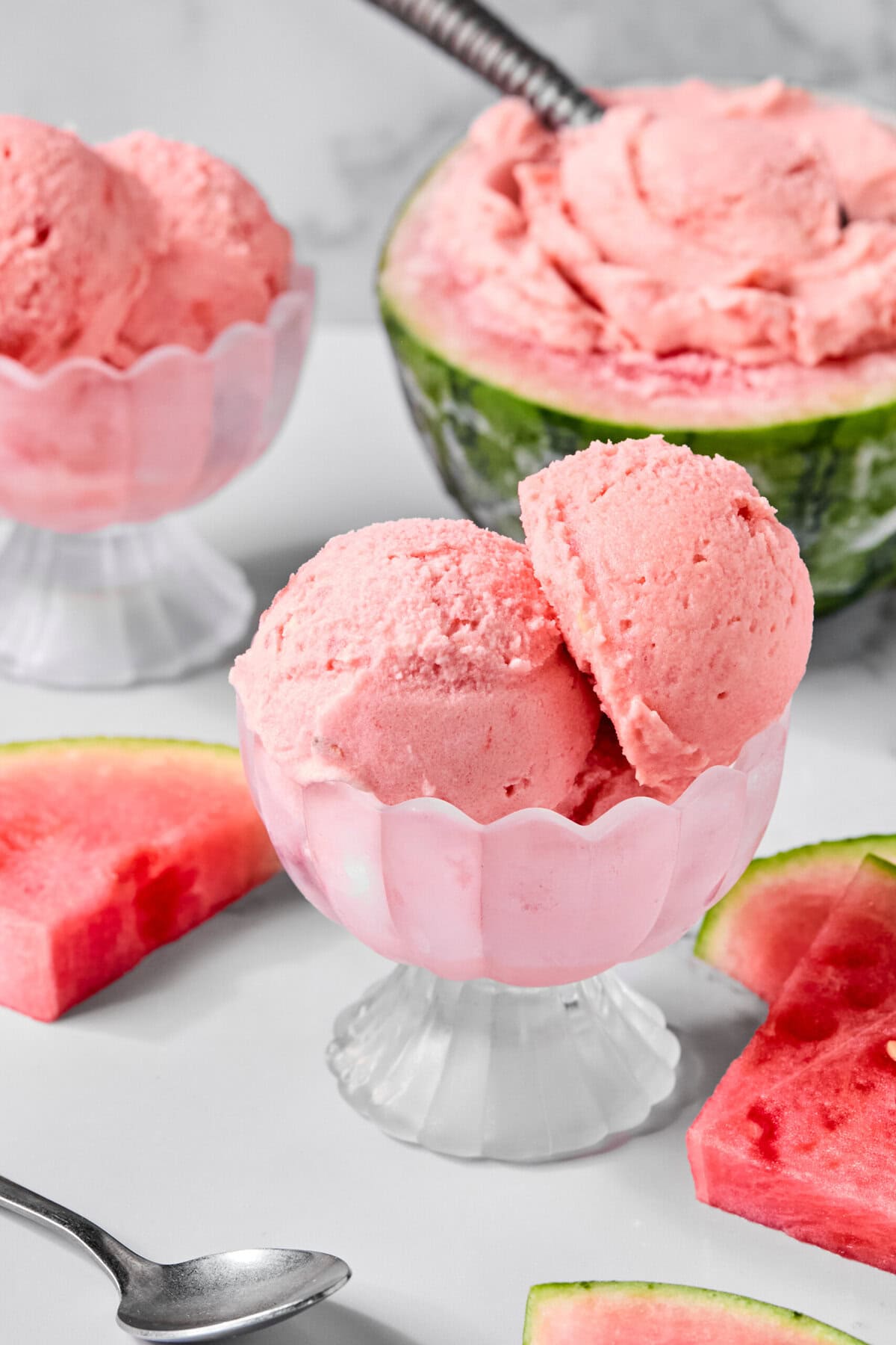 Two scoops of pink watermelon ice cream in a glass dish, with watermelon slices and a watermelon half filled with ice cream in the background. A spoon sits on the table, evoking the refreshing flair of a trending TikTok watermelon ice cream recipe.