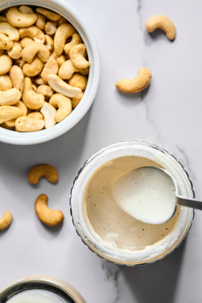 A bowl of cashews sits beside a jar of creamy white sauce with a spoon inside, evoking the fresh, homemade vibes seen in trending TikTok watermelon ice cream recipe videos. Several loose cashews are scattered on a white marble surface.