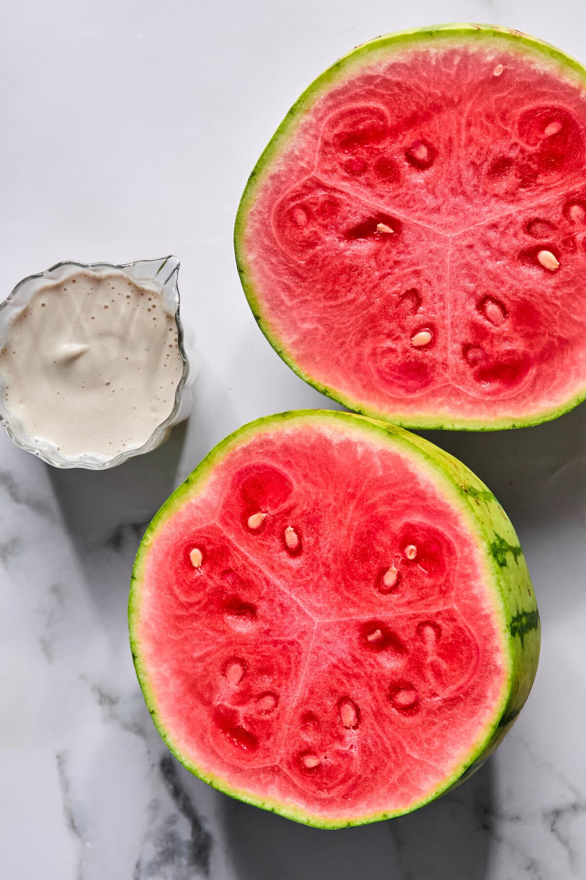 Two halves of a ripe watermelon with bright red flesh are placed on a marble surface next to a small bowl of creamy white sauce, reminiscent of the viral tiktok watermelon ice cream recipe.