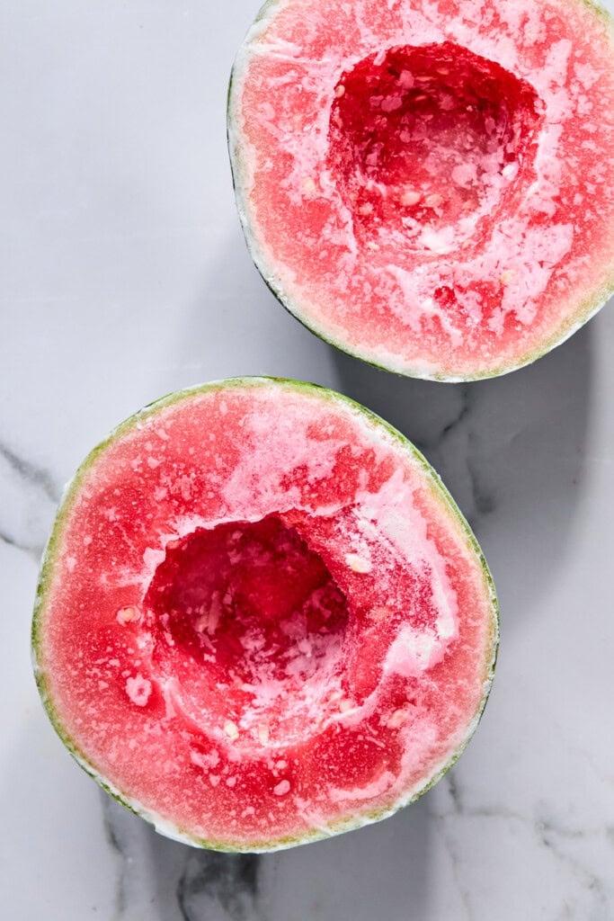 Two halves of a watermelon with their centers scooped out, exposing red flesh and white rind, placed on a white marble surface-perfect preparation for trying the viral TikTok watermelon ice cream recipe.