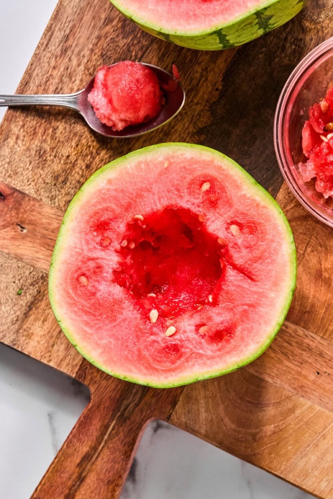A halved watermelon with its center scooped out sits on a wooden board, ready to become the star of your next TikTok watermelon ice cream recipe. A spoon with watermelon flesh and a small bowl of scooped watermelon are nearby, perfect for mixing or serving this trending treat.
