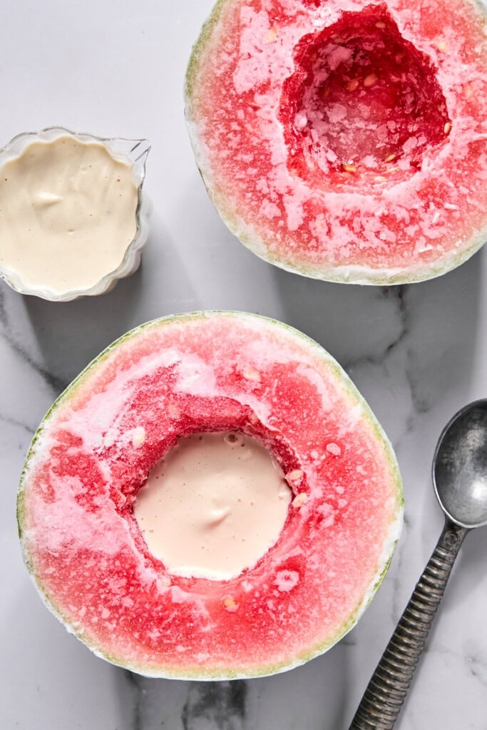 A hollowed-out watermelon half filled with creamy sauce sits next to a matching empty half, a small bowl of sauce, and a metal spoon on a marble surface-perfect for trying out the latest TikTok watermelon ice cream recipe.