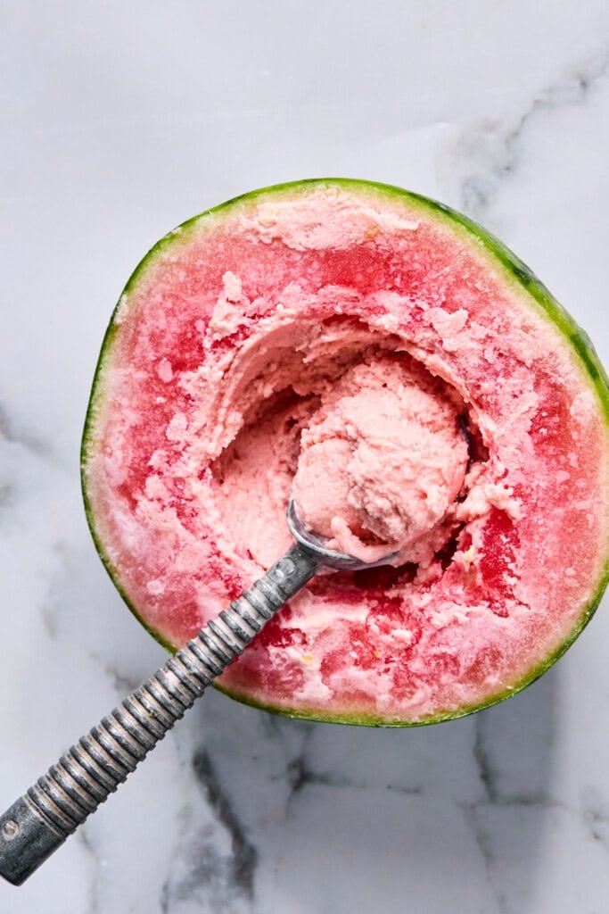 A metal spoon scoops pink ice cream from a hollowed-out half of a watermelon, placed on a white marble surface-perfect for recreating that viral TikTok watermelon ice cream recipe.