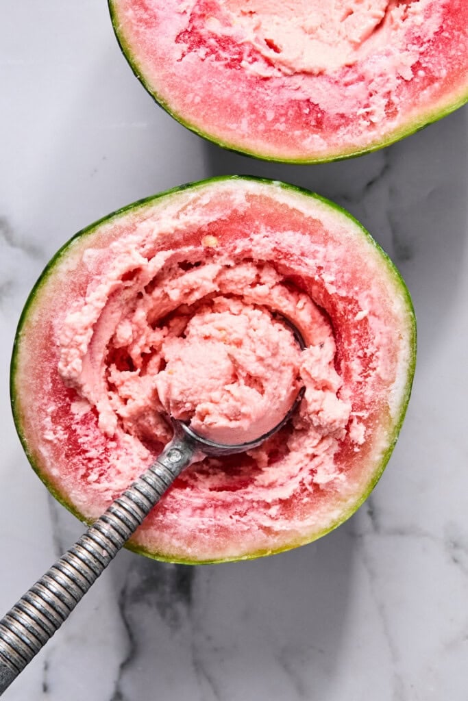 A metal ice cream scoop sits in a halved watermelon filled with pink watermelon ice cream, inspired by the viral TikTok watermelon ice cream recipe, on a white marble surface.
