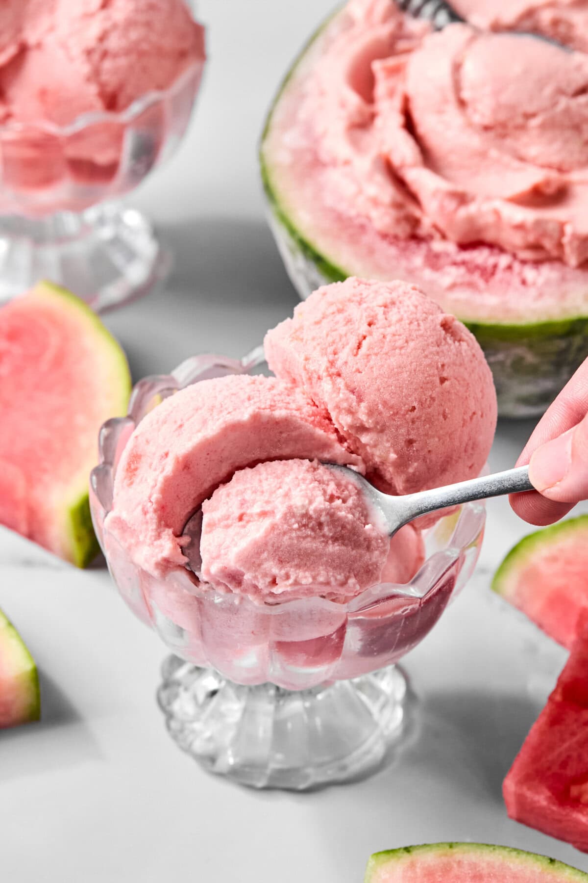 A hand holding a spoon serves pink watermelon sorbet into a glass dessert bowl, inspired by the viral tiktok watermelon ice cream recipe, with watermelon slices and another bowl of sorbet in the background.