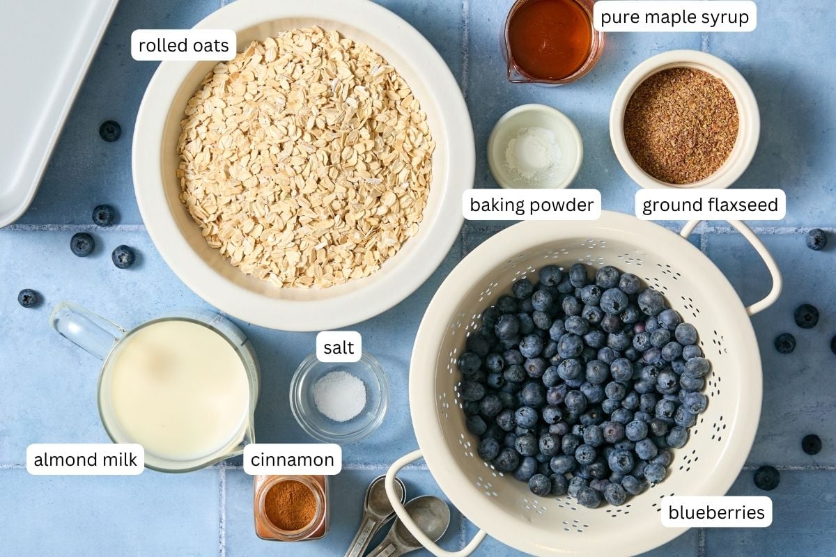 A flat-lay of labeled ingredients for blueberry baked oats: rolled oats, blueberries, almond milk, cinnamon, salt, baking powder, ground flaxseed, and pure maple syrup on a blue surface.