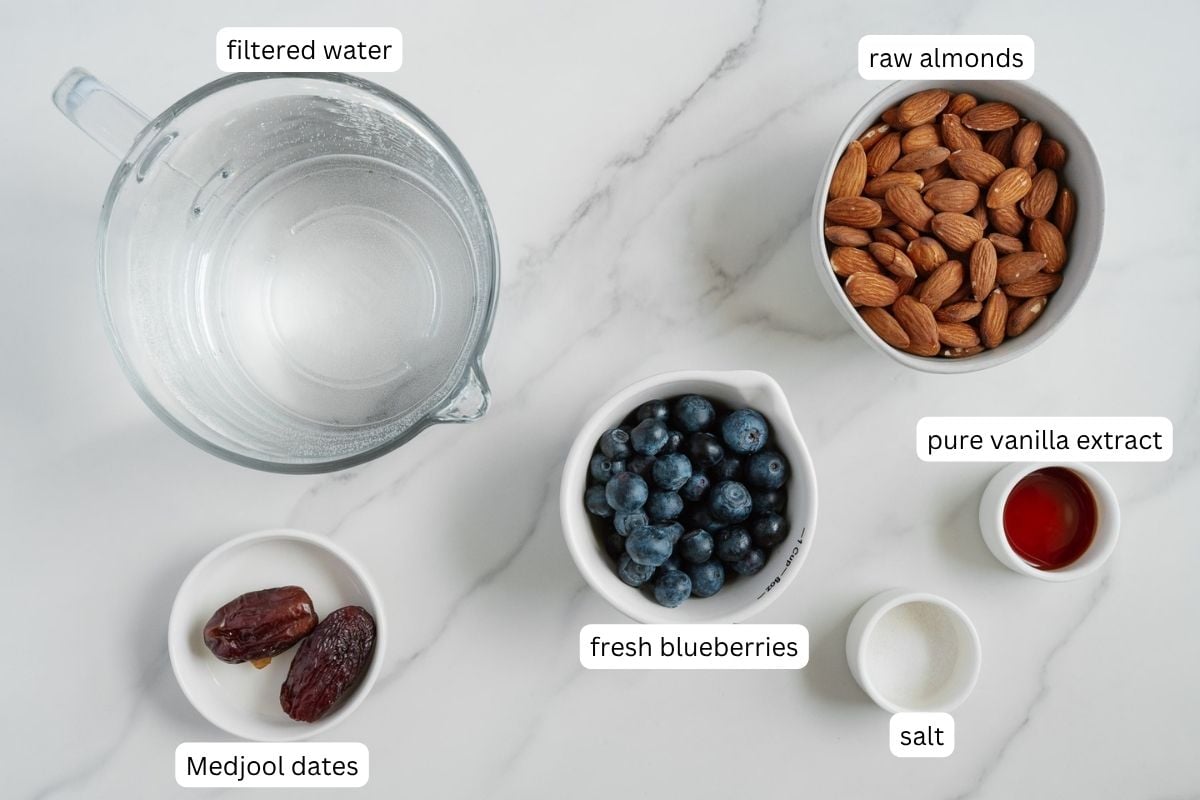 A glass measuring cup of filtered water, a bowl of raw almonds, a small bowl of fresh blueberries, two Medjool dates for naturally sweetened flavor, a small glass of pure vanilla extract, and a pinch of salt-all labeled on a white marble surface-come together to create vibrant blueberry almond milk that's completely dairy-free.
