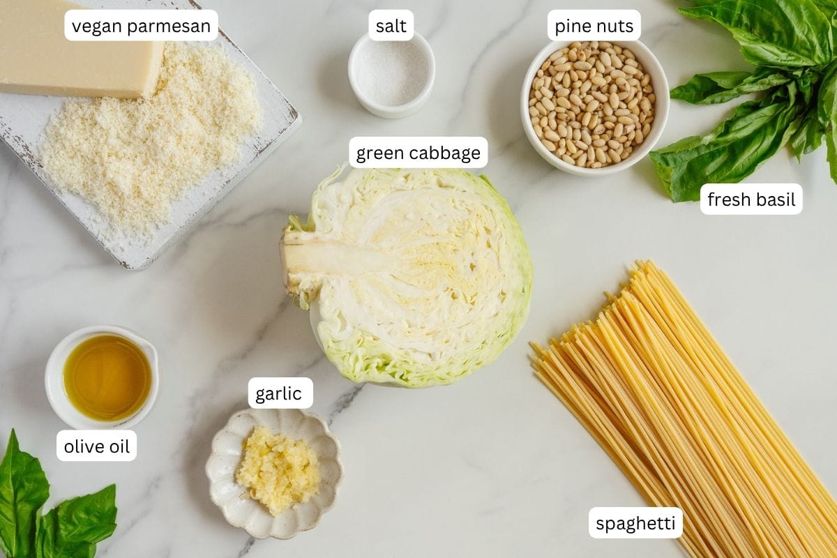 Ingredients for a caramelized cabbage pasta dish: half a green cabbage, dry spaghetti, vegan parmesan, pine nuts, fresh basil, salt, garlic, and olive oil arranged on a white surface, each labeled.