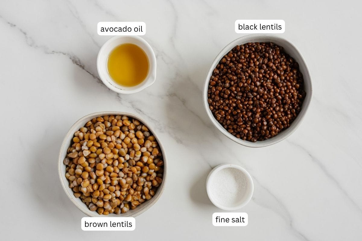 Four bowls on a white surface containing labeled ingredients: avocado oil, black lentils, brown lentils, and fine salt—everything you need to make perfectly crispy lentils.