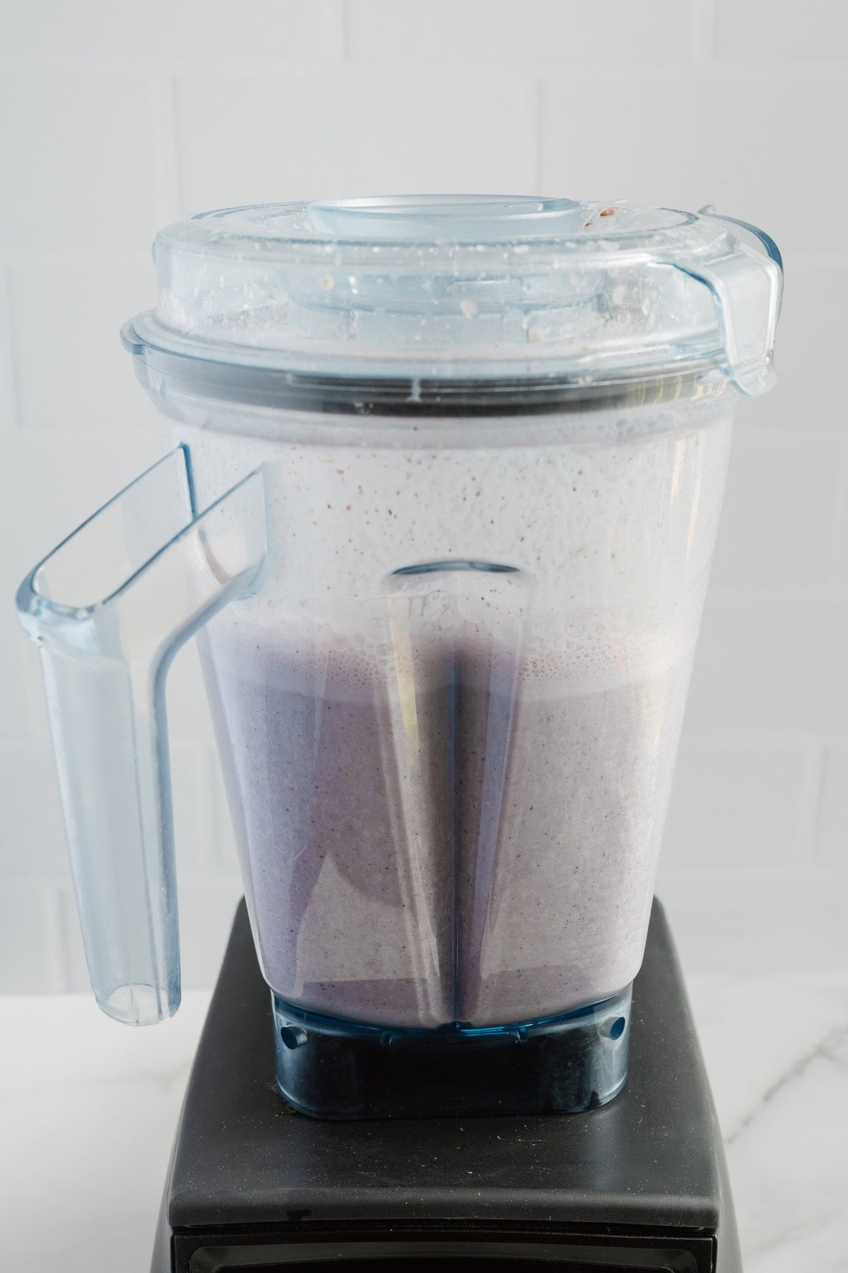 A blender filled with a light purple, naturally sweetened Blueberry Almond Milk smoothie mixture sits on a black base against a white tiled background.