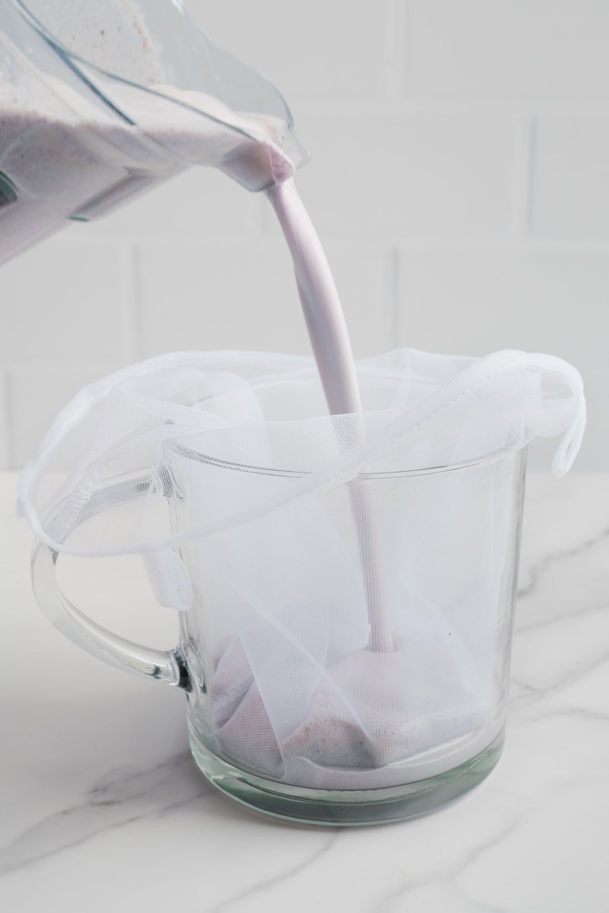 A blender is pouring a naturally sweetened, dairy-free blueberry almond milk-a light purple liquid-through a white nut milk bag into a clear glass pitcher on a white countertop.
