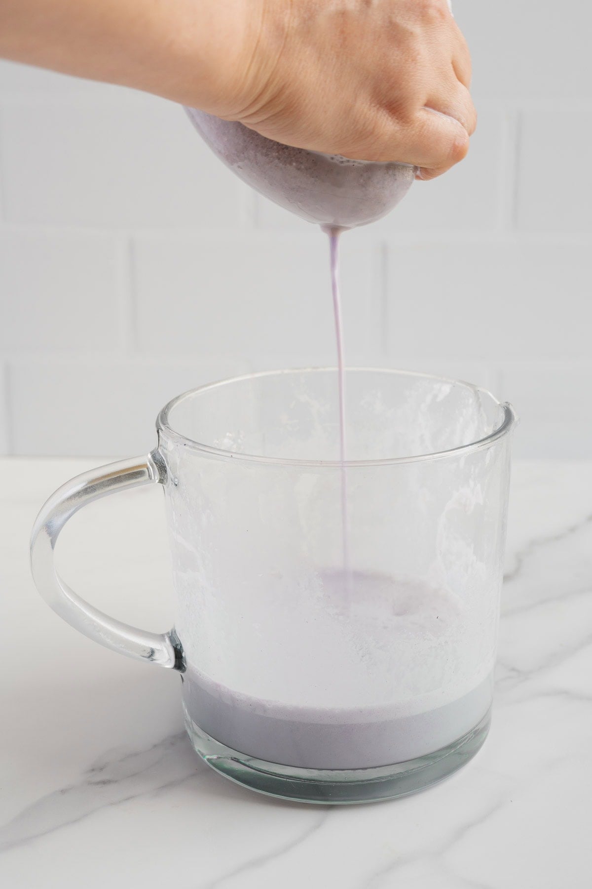 A hand squeezes a nut milk bag, straining a vibrant purple liquid-Blueberry Almond Milk-into a clear glass pitcher on a white countertop with a tiled background. This dairy-free beverage is naturally sweetened, offering a delicious and wholesome alternative to traditional milk.