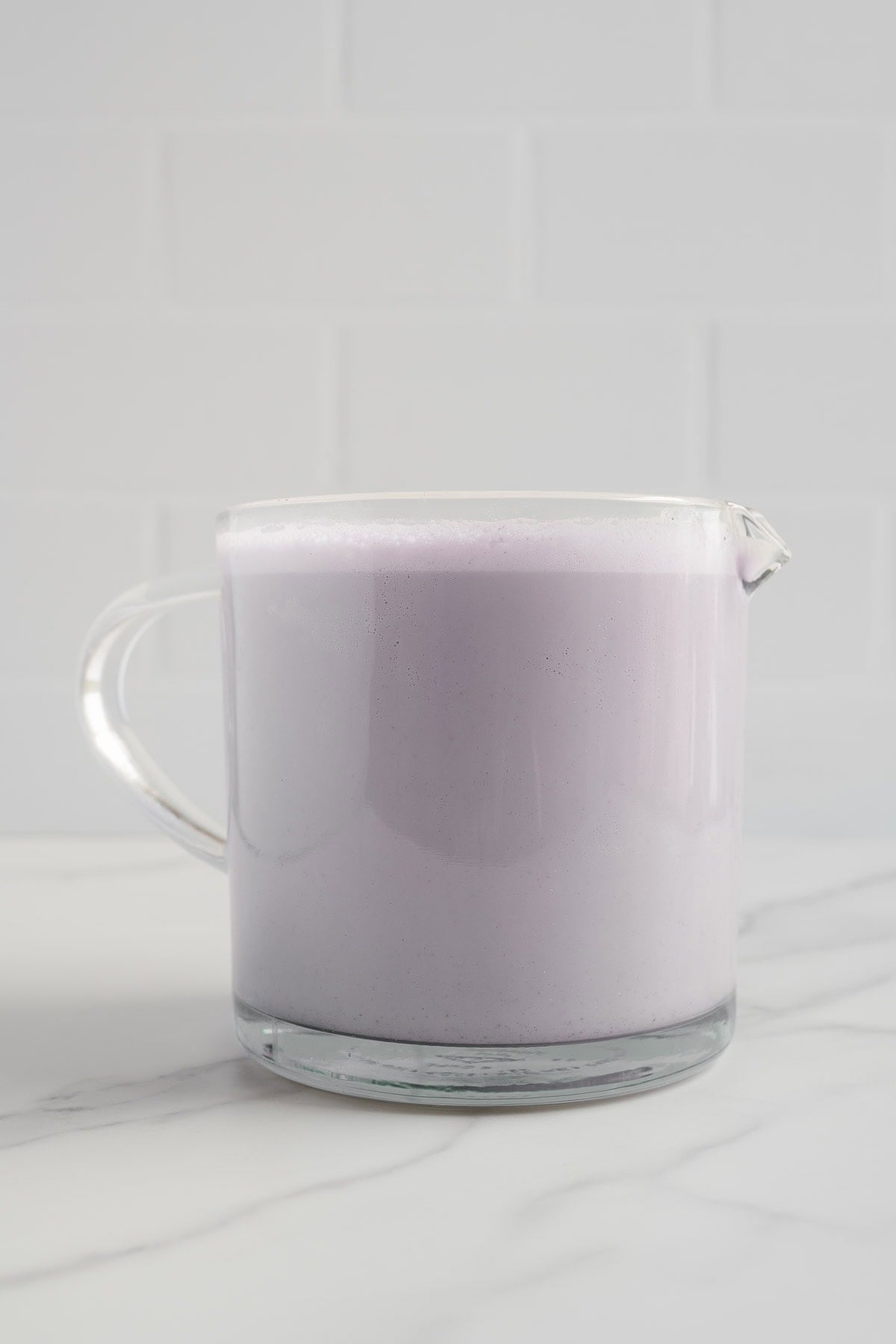 A clear glass pitcher filled with light purple, naturally sweetened blueberry almond milk sits on a white marble surface, with a white tiled wall in the background. This refreshing beverage is completely dairy-free.