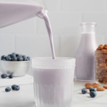 A glass is being filled with naturally sweetened blueberry almond milk, with fresh blueberries, almonds, and a bottle of the same dairy-free milk in the background on a white surface.