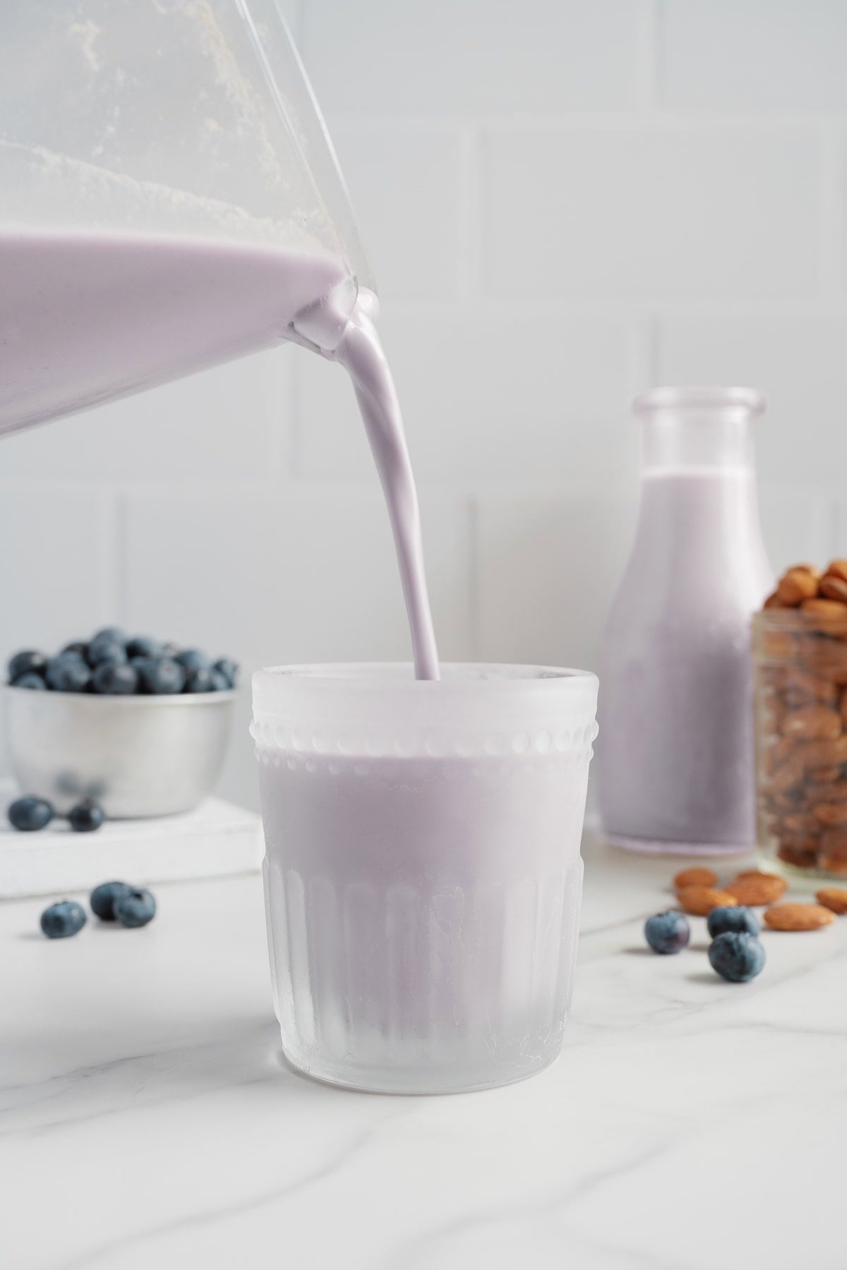 A glass is being filled with naturally sweetened blueberry almond milk, with fresh blueberries, almonds, and a bottle of the same dairy-free milk in the background on a white surface.