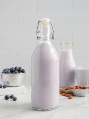 A glass bottle filled with naturally sweetened blueberry almond milk stands on a white counter, with a bowl of blueberries, a tray of almonds, and another bottle and glass of the same dairy-free milk in the background.