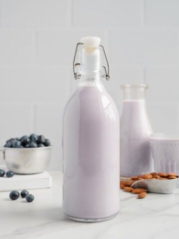 A glass bottle filled with naturally sweetened blueberry almond milk stands on a white counter, with a bowl of blueberries, a tray of almonds, and another bottle and glass of the same dairy-free milk in the background.