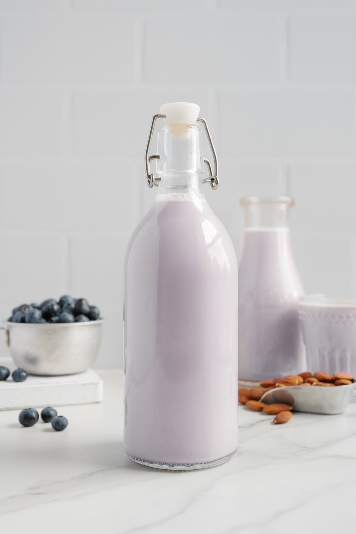 A glass bottle filled with naturally sweetened blueberry almond milk stands on a white counter, with a bowl of blueberries, a tray of almonds, and another bottle and glass of the same dairy-free milk in the background.