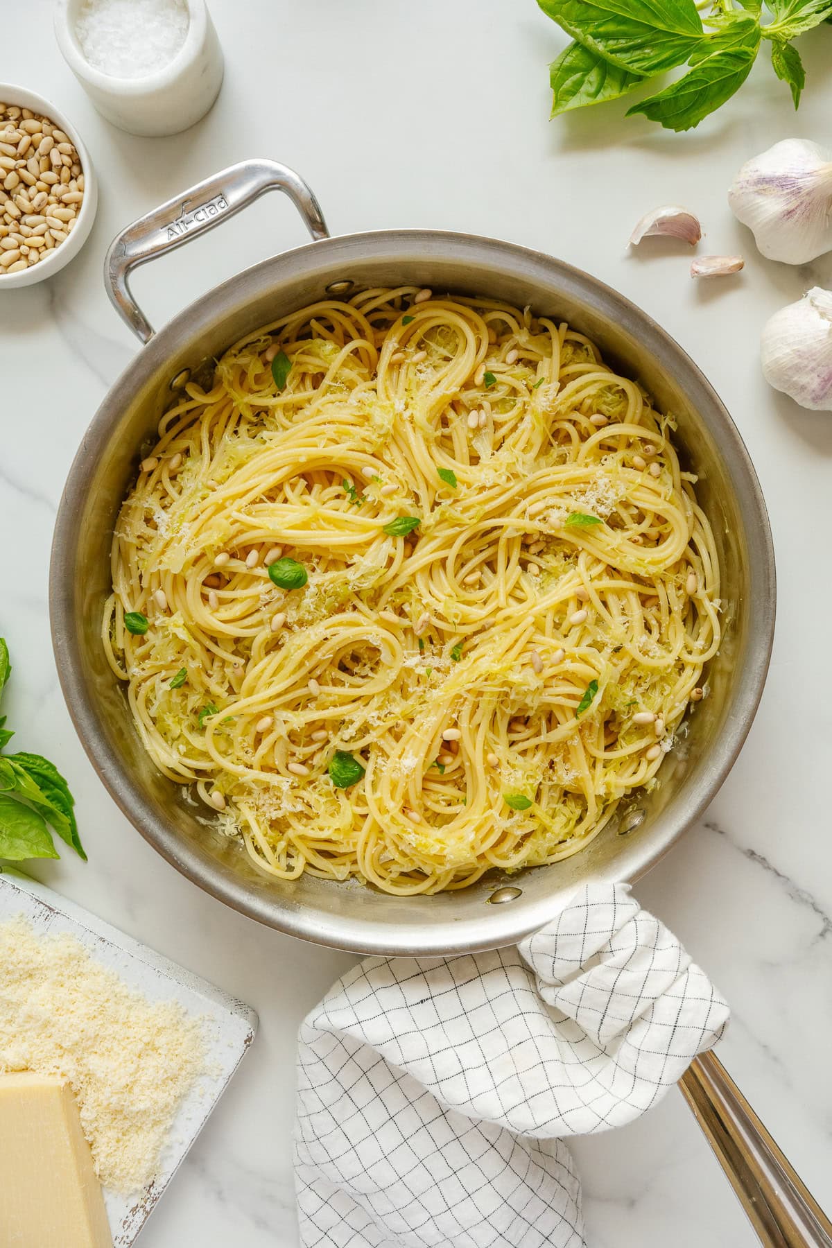 A skillet of cooked spaghetti pasta with grated cheese, pine nuts, and fresh basil, surrounded by garlic, parmesan, salt, and basil leaves on a white marble surface.