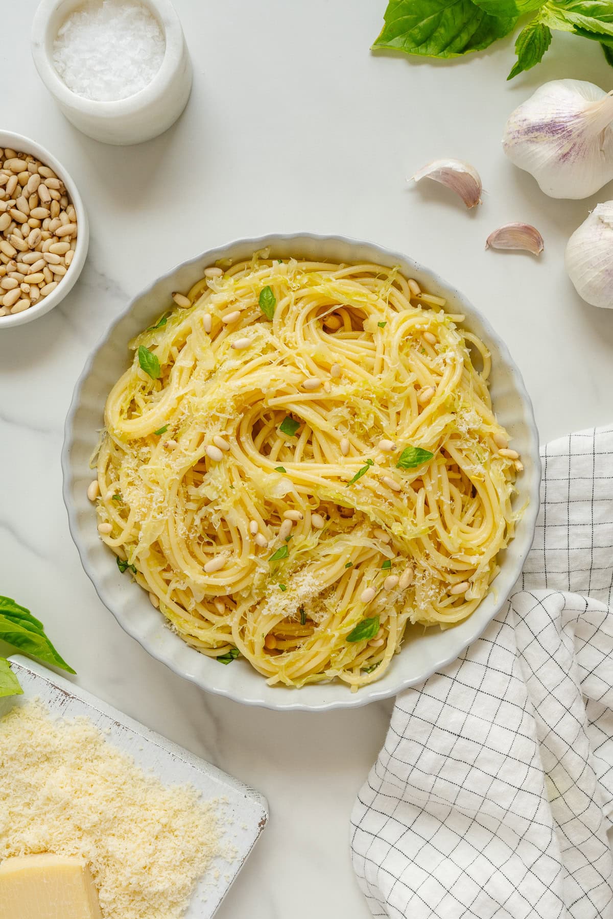 A bowl of spaghetti garnished with grated cheese, basil, and pine nuts sits on a marble surface next to garlic, parmesan, a dish of salt, pine nuts, basil leaves, and a checkered cloth.