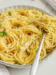 A plate of spaghetti with pine nuts, grated cheese, and basil, with a fork resting in the pasta. Garlic, pine nuts, and cheese are in the background on a white surface.