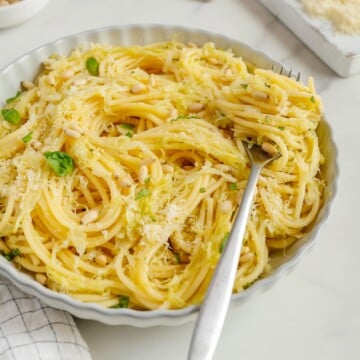 A plate of spaghetti with pine nuts, grated cheese, and basil, with a fork resting in the pasta. Garlic, pine nuts, and cheese are in the background on a white surface.