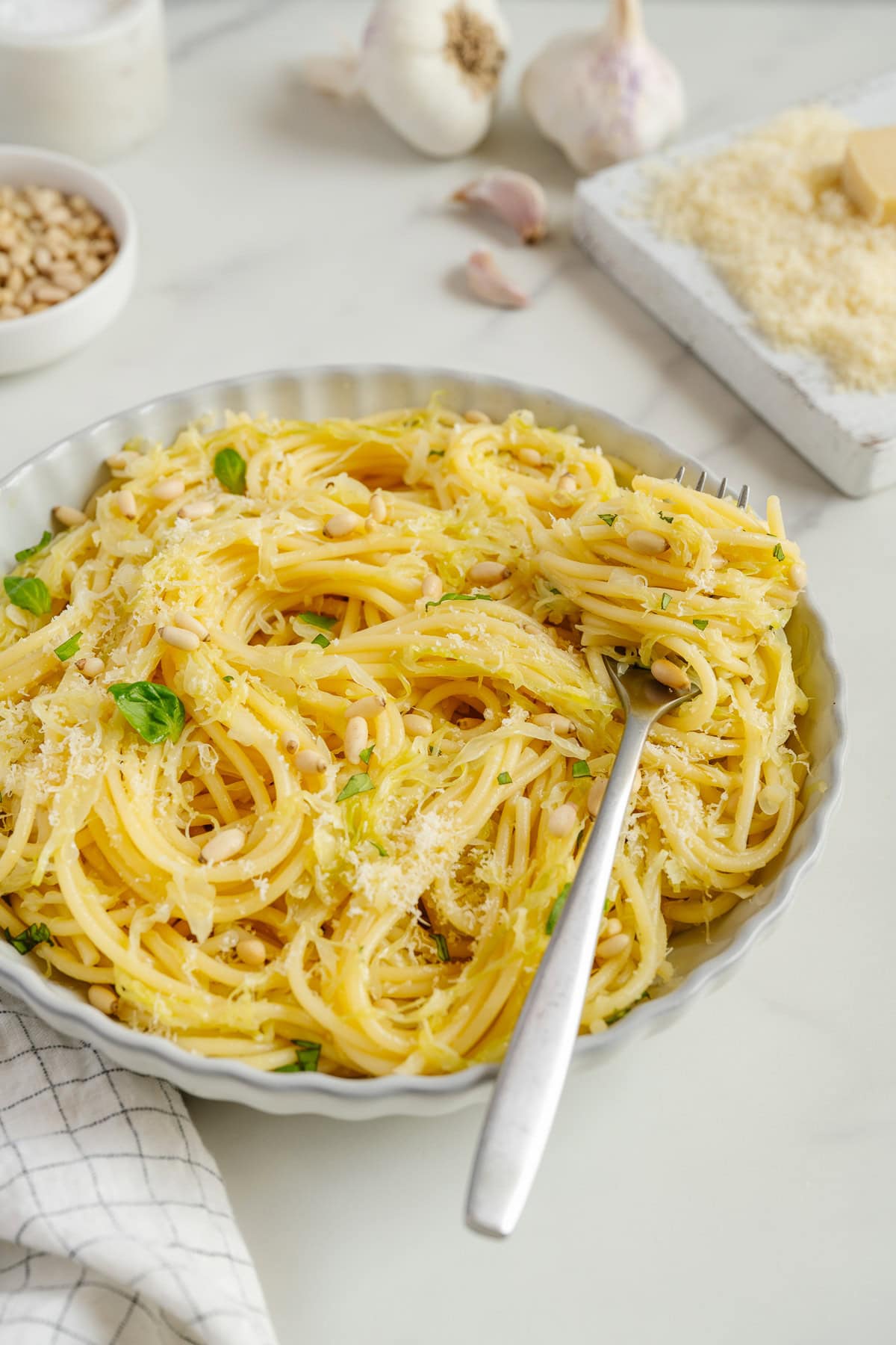 A plate of spaghetti with pine nuts, grated cheese, and basil, with a fork resting in the pasta. Garlic, pine nuts, and cheese are in the background on a white surface.