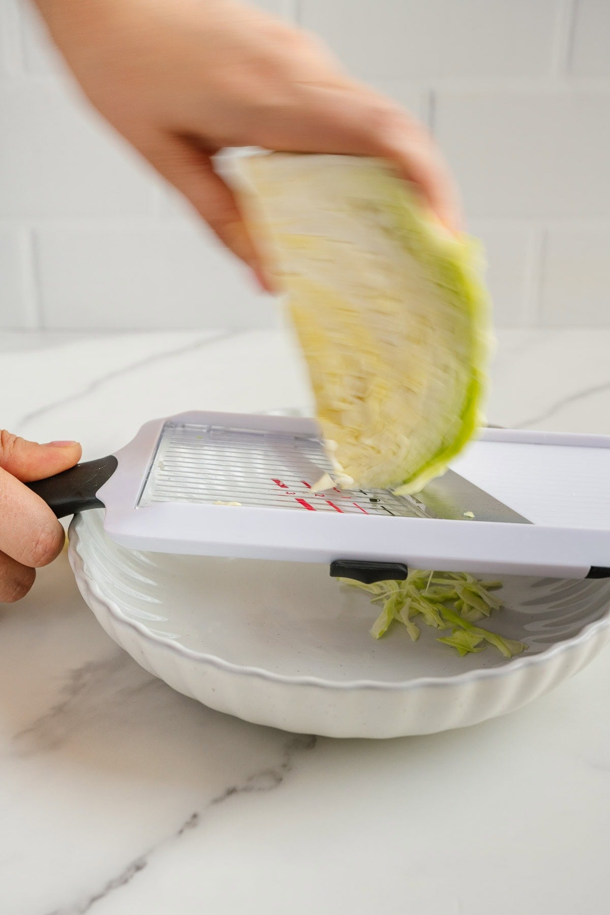 A hand slices cabbage with a white mandoline slicer over a white bowl, with thin cabbage shreds collecting below on a white countertop.