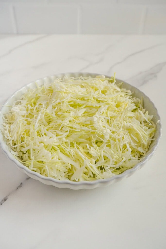 A round white dish filled with finely shredded cabbage sits on a white marble countertop with a light gray vein pattern.