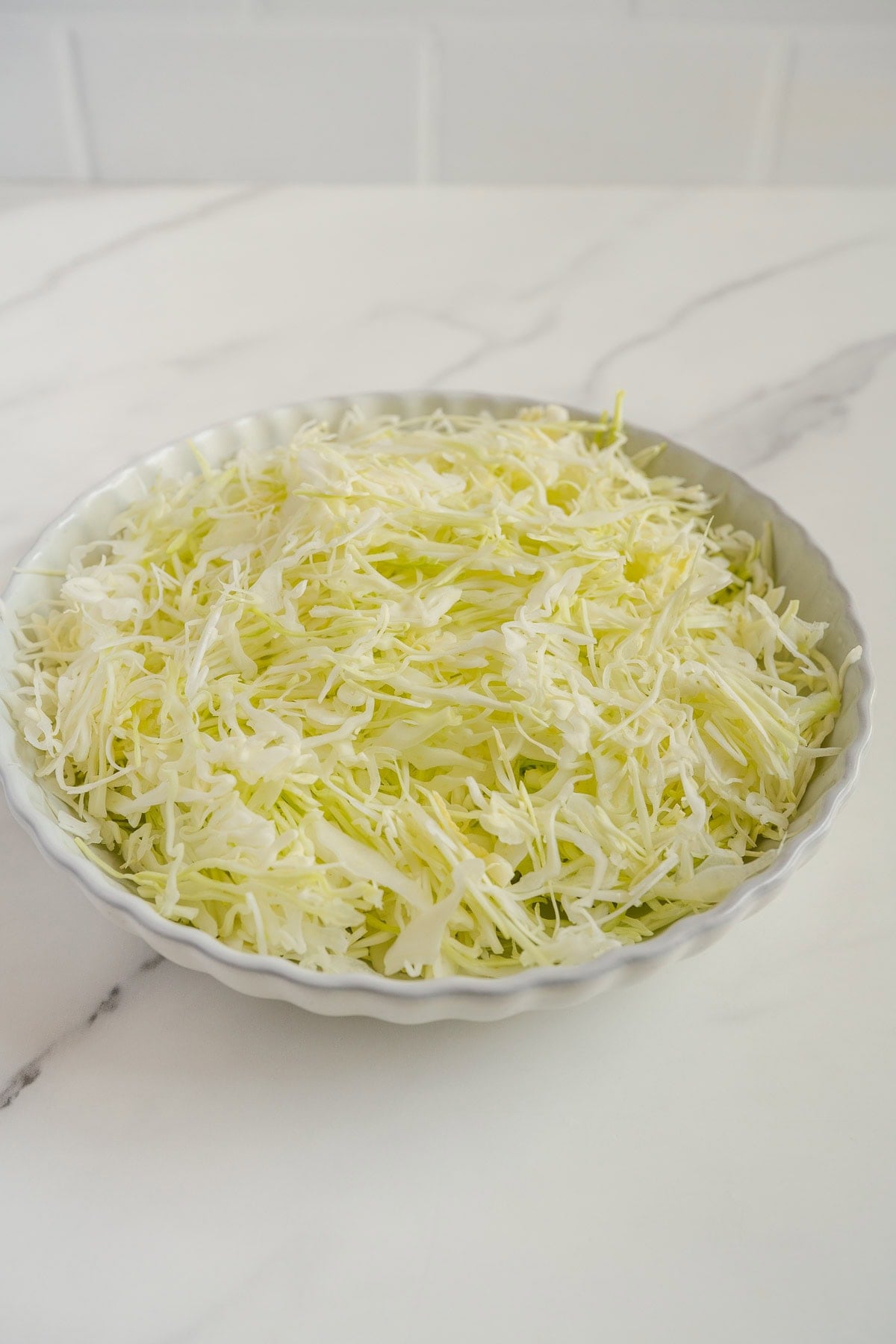 A round white dish filled with finely shredded cabbage sits on a white marble countertop with a light gray vein pattern.