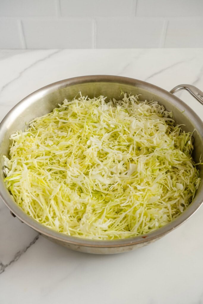 A stainless steel pan filled with finely shredded raw cabbage sits on a white marble countertop with a white tiled background.