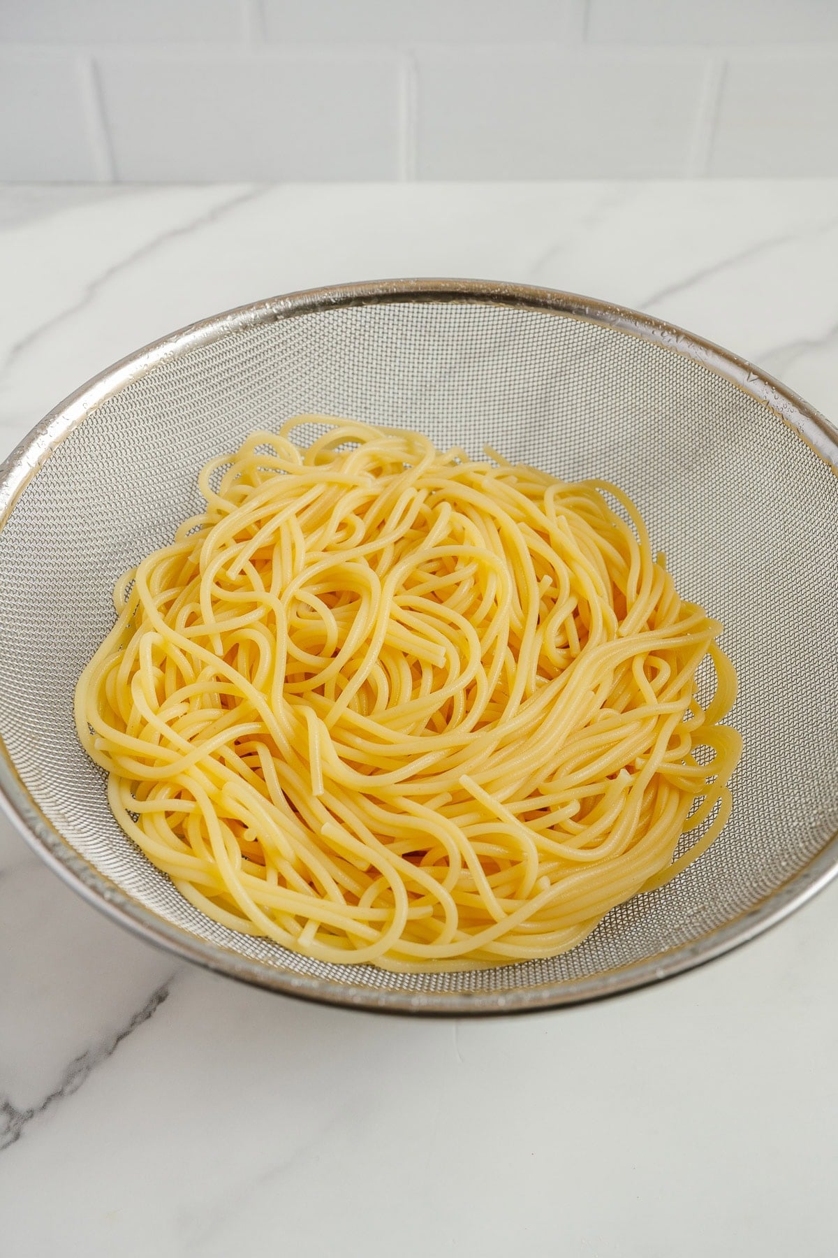A metal strainer filled with cooked spaghetti sits on a white marble countertop, with a white tiled wall in the background.