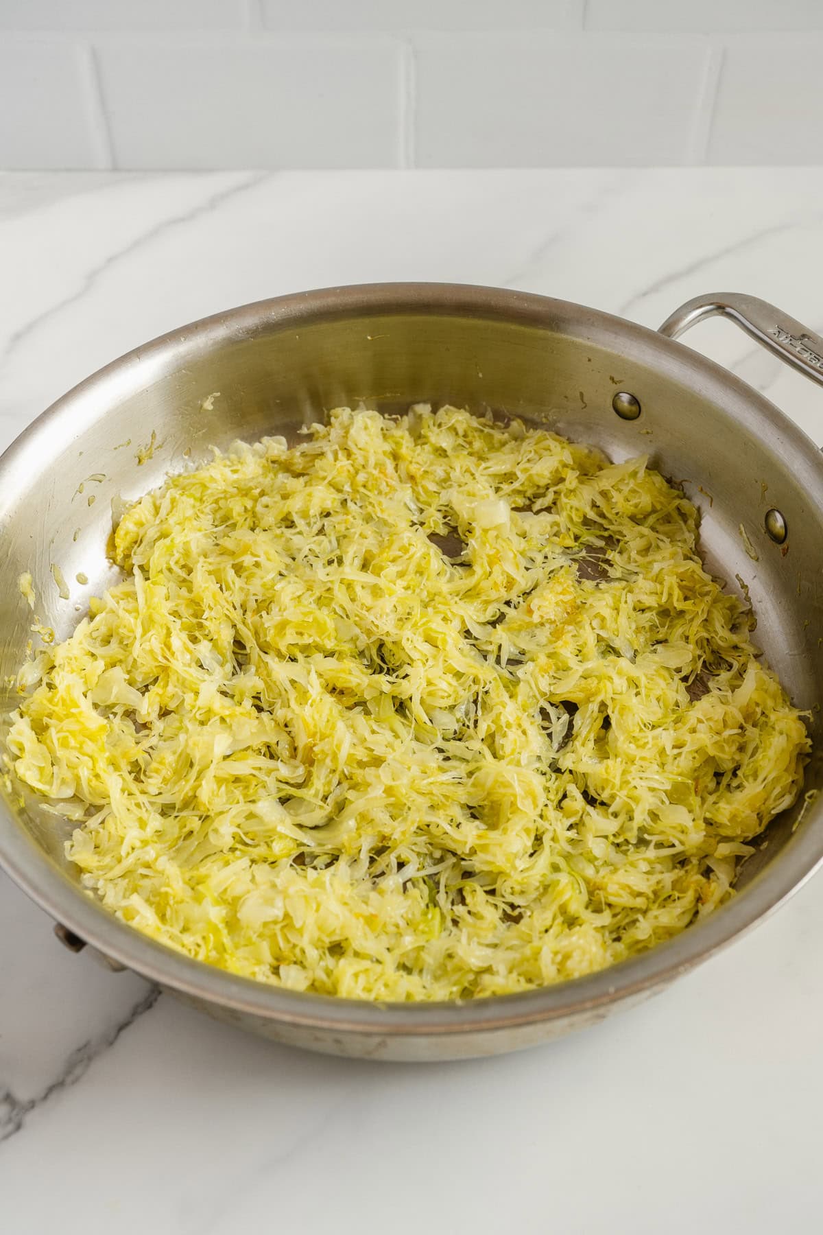 Shredded sautéed cabbage in a stainless steel skillet sits on a white marble countertop, with light browning visible and a tiled white backsplash in the background.