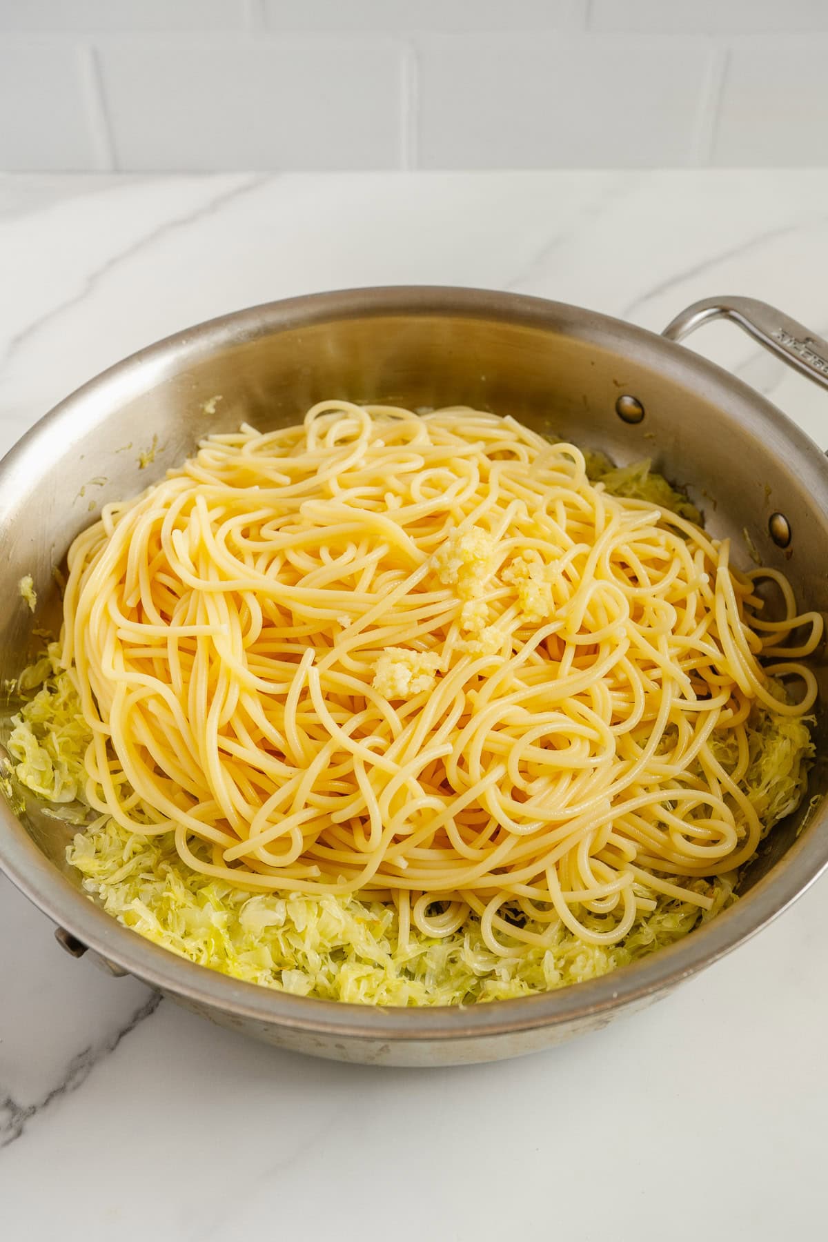 A metal pan filled with cooked spaghetti, sautéed shredded cabbage, and minced garlic on a white marble surface.