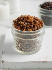 A small glass jar filled with crispy, golden brown lentils sits on a white wooden board, with another jar of crispy lentils, darker in color, blurred in the background.