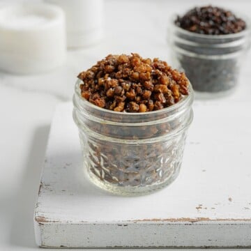A small glass jar filled with crispy, golden brown lentils sits on a white wooden board, with another jar of crispy lentils, darker in color, blurred in the background.