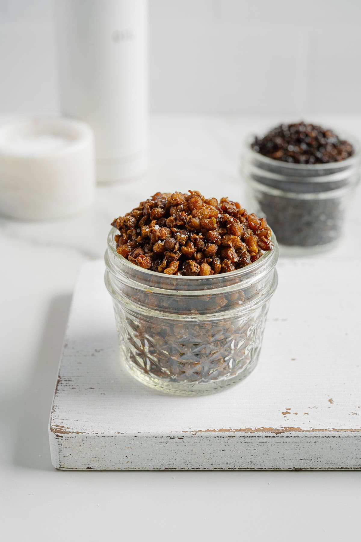 A small glass jar filled with crispy, golden brown lentils sits on a white wooden board, with another jar of crispy lentils, darker in color, blurred in the background.