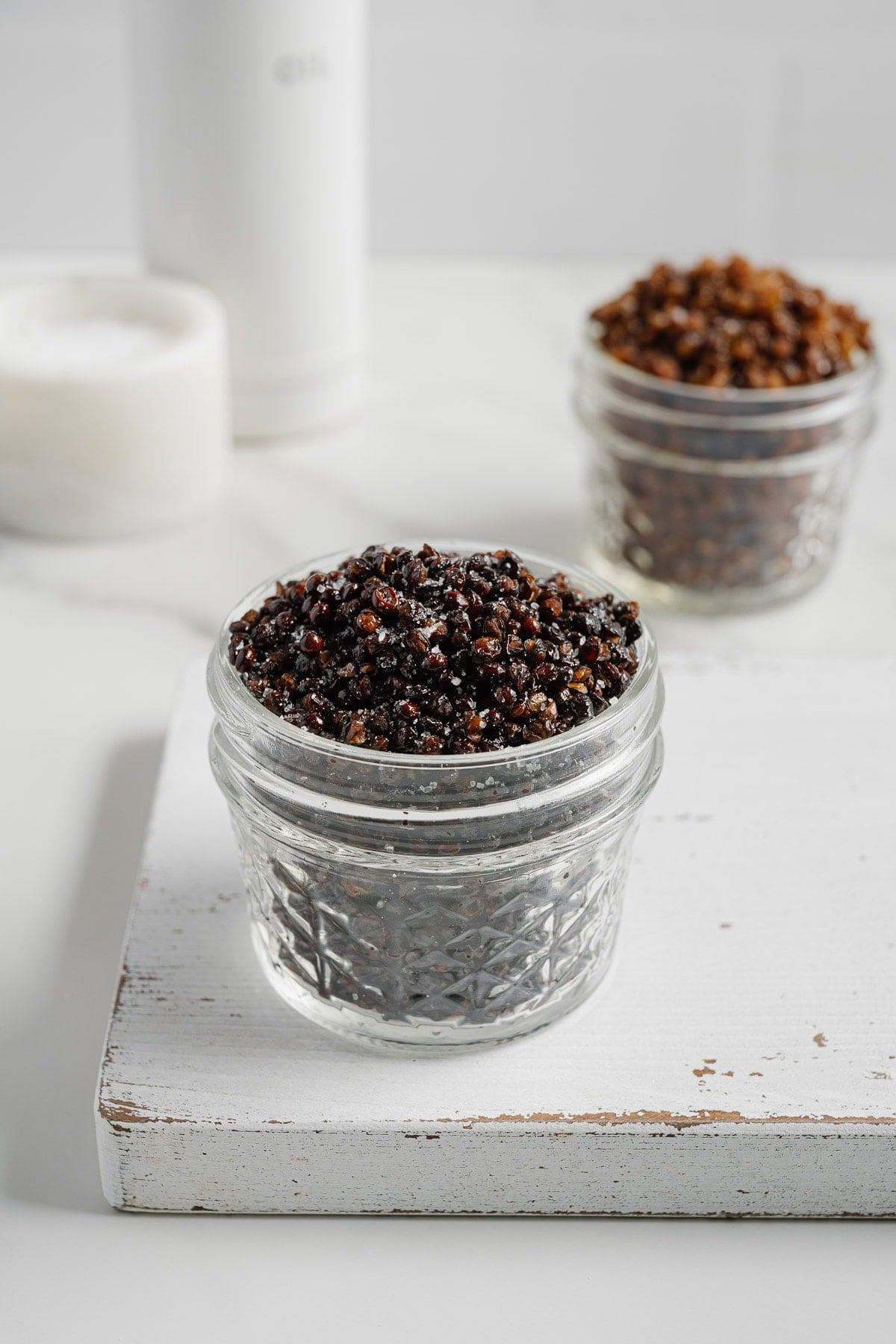 A small glass jar filled with crispy lentils sits on a white wooden board, accompanied by another similar jar and a white container in the background.