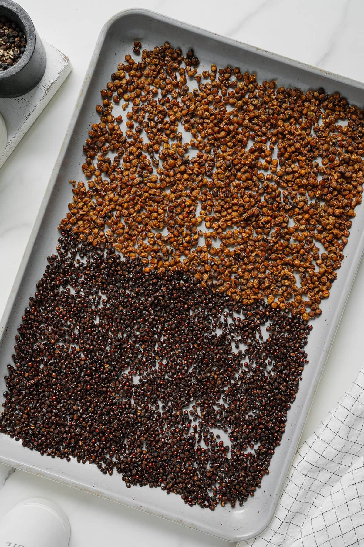A baking sheet on a white surface holds two sections of roasted chickpeas—lighter brown on top and darker brown on the bottom—reminiscent of the inviting texture of crispy lentils. A mortar and pestle and a white cup are nearby.